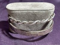 Two sterling silver bracelets displayed on a gray velvet stand against a purple fabric background. The front bracelet has a hammered finish, and the other features a twisted rope design.
