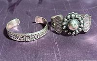 Two sterling silver cuff bracelets side by side on purple fabric. The left bracelet has a floral design; the right bracelet has Native American style arrowhead and beaded dome embellishments.