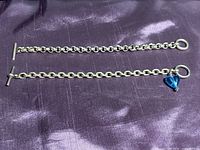 Two sterling silver rolo chain bracelets placed side by side on a purple fabric background, one plain and one with a blue glass heart charm.