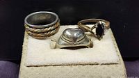 Three sterling silver rings displayed on a beige ring holder against a black background. Left ring has twisted pattern, center is a heart-shaped solid silver ring, right ring has a black marquise stone.