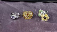 Three sterling silver rings displayed on a dark fabric. Left is a silver ring with purple and clear stones, center is a gold plated band ring with multiple gemstones, right is a flower-shaped ring with yellow and green stones.