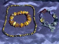 Three pieces of jewelry on purple fabric: a round cats eye bead necklace with sterling silver S clasp; a stretch amber chip bracelet; a sterling silver bracelet with amethyst, citrine, red glass beads and toggle clasp.