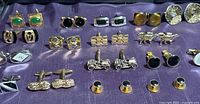 Set of 15 cufflinks and buttons displayed on purple cloth surface including designs with stones, enamel, and textured metal finishes.