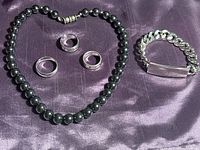 Lot contents displayed on dark purple fabric: hematite necklace, three stainless steel rings, and a chain link bracelet with 'Bill' engraved name tag.