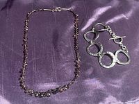 Both items laid out side by side on purple satin fabric. Bracelet with large hammered sterling silver circle links and toggle clasp. Necklace is a delicate chain with small garnet drops.