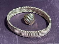 Sterling silver flexible mesh bangle bracelet laid in an oval shape alongside a sterling silver conch wave ring on purple fabric background.