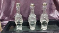 Three clear crystal bottles with sterling silver screw-on tops set against a purple backdrop on a black tray.