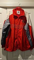 Front view of red, white, and gray Buccaneers windbreaker jacket hung on hanger against white door.