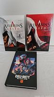Three books displayed standing: two Assassin's Creed novels and one Call of Duty Black Ops 4 hardcover graphic novel.