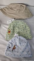 Photo showing three baby hats: beige bucket hat, light green Winnie the Pooh beanie, light blue Tigger beanie