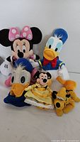 Photo showing five assorted Disney plush toys including Minnie Mouse, two Donald Ducks in different outfits, a small Mickey Mouse in yellow dress, and a Pluto plush toy.