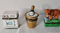Front view of three vintage hinged trinket boxes: stove, ice bucket with bottle, bear on grass base
