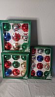 Three unopened boxed sets of colorful Rauch glass Christmas ornaments arranged on a table.