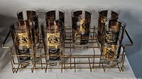Seven tall glass tumblers in a gold metal wire caddy with black and gold wonder of the world designs on clear glass.