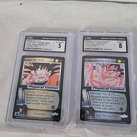 Photo of two Dragonball Z trading cards graded and encapsulated by CGC, featuring images of Goku in combat moves titled 'Physical Combat'. One card graded 5, the other graded 8.
