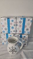 Stack of four boxes with snowflake pattern; one mug in plastic wrap placed in front