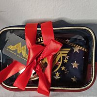 Clear plastic case with gold edging and red ribbon revealing Wonder Woman cosmetic bag set inside.