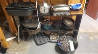 Shelved assortment showing electric grill, stainless pot, green pot, speckled roasters, muffin tins, whisks