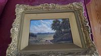 Entire framed painting on burgundy surface showing ornate gilt frame and landscape scene