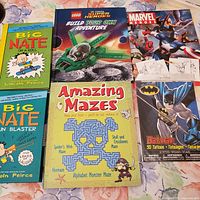 Top view of six children's books arranged overlapping on a floral fabric background. Books visible include Big Nate On A Roll, Big Nate Fun Blaster, Lego DC Comics Super Heroes Build Your Own Adventure, Marvel Heroes, Batman 50 Tattoos, and Amazing Mazes.