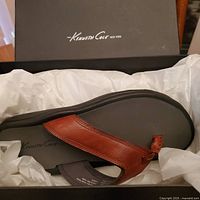 Left angled view of Kenneth Cole sandal inside black box with white tissue paper, showing brown leather strap and black footbed with logo.