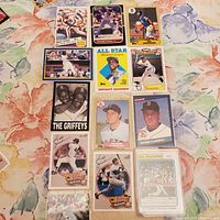 Photo showing an assortment of 14 vintage Major League Baseball cards featuring players including Roger Clemens, Dwight Gooden, Ken Griffey Jr., Don Mattingly, and others arranged on a floral fabric background.