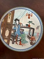 Front view of the porcelain decorative plate showing a seated Asian woman in traditional attire inside a room with bonsai and other decorations, gilt edge border visible.