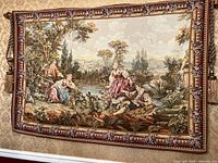 Full view of large pastoral scene tapestry hung on wall showing detailed scene and floral border with attached rod and tassel.