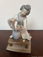 Porcelain figurine showing East Asian woman arranging pink flowers in a vase on a small brown table. Figure dressed in traditional white and blue-gray robe.