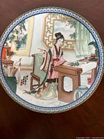 Top side of 8-inch decorative porcelain plate with traditional Oriental painting of a woman in a room with furniture and nature details, multicolored design with blue and red border.