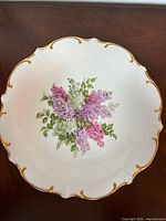 Top view of the porcelain plate showing floral design with purple, pink, white flowers and green leaves with gold-trimmed scalloped edge.