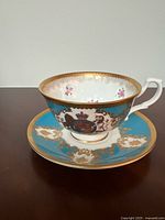 Photo shows the porcelain teacup with a gold and blue royal crest design along with pink floral interior details, sitting on the matching saucer.