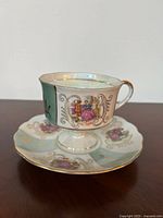 Porcelain cup and saucer set displayed with visible Victorian era scene and gold decorative accents, showing front of cup and saucer.