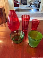 Photo of all five glass pieces: three red vases of various shapes and two green bowls on a table in a home setting