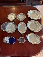 Full collection of silver serving dishes arranged on a wood table showing multiple trays, bowls, and glass inserts.