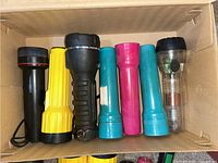 Seven assorted cylindrical flashlights standing upright in a cardboard box