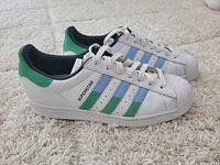 Side view showing white leather Adidas Superstar shoes with green and blue stripes, shell toes, and white laces on carpet.