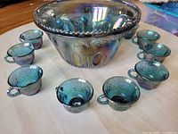 Blue iridescent punch bowl with 7 punch glasses displayed around it on table, showing grape leaf pattern and iridescent finish