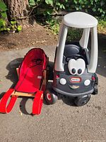 Photo showing both the wooden Kinder sleigh with red fabric seat and the Little Tikes police car parked side by side on pavement outdoors.