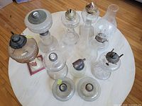 Overview of nine kerosene lamps and various glass chimneys, some lamps with metal burners attached and some chimneys detached.