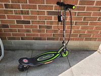 Side view of Razor Powercore E90 electric scooter parked against a brick wall, showing black deck with green details and rear wheel.