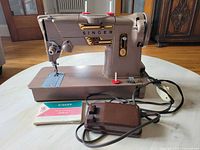 Front side view showing Singer logo, stitch length and tension controls, two red spool holders on top, and power cord with foot pedal.