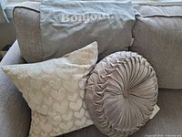 Three throw pillows on couch: rectangular gray 'Bonjour' pillow cover, square cream heart pattern pillow, and round gray tufted pillow.