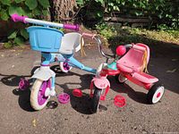 Photo showing two children's tricycles on pavement, one blue and white with purple accents and a blue rear basket, and one pink with red pedals and seat.