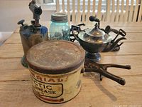 Photo showing a side view of the lot items on a wooden table including tin, burner, canning jar, boot jack and apple peelers.