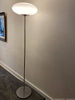 Full view of the silver chrome metal floor lamp with opaque white glass shade lit and standing against a wall.
