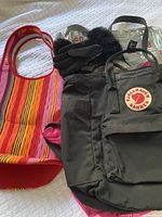 Photo showing a striped neoprene tote bag with red handles, black fur-trimmed gloves resting on a dark Fjällräven Kånken backpack with front pocket and logo patch.