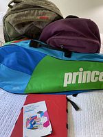 Photo showing two backpacks (one beige, one purple) and a large multicolored sports bag with 'prince' branding, clothes folding board below them.
