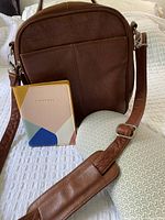 Front view of the tan leather crossbody bag with adjustable strap, multicolor geometric passport holder and fabric travel pillow on a white textured surface