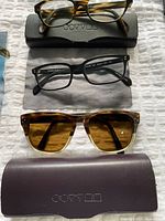 Three pairs of Oliver Peoples glasses, two eyeglasses and one sunglasses, displayed on a white textured surface with their respective branded cases and cleaning cloths.
